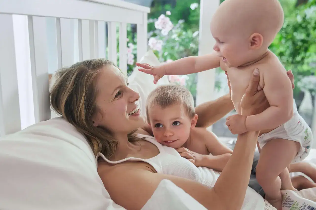 Gl&uuml;ckliche Familie im Bett. Ein Vater h&auml;lt sein Baby hoch, w&auml;hrend die Mutter und das &auml;ltere Kind zuschauen. Ein sch&ouml;nes Bild f&uuml;r den Vaterschaftsurlaub in Deutschland.