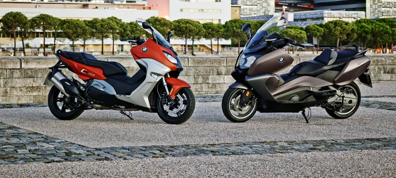 Dwa skutery BMW C 650 GT w kolorach pomarańczowym i brązowym stoją obok siebie na tle miejskiego krajobrazu.
