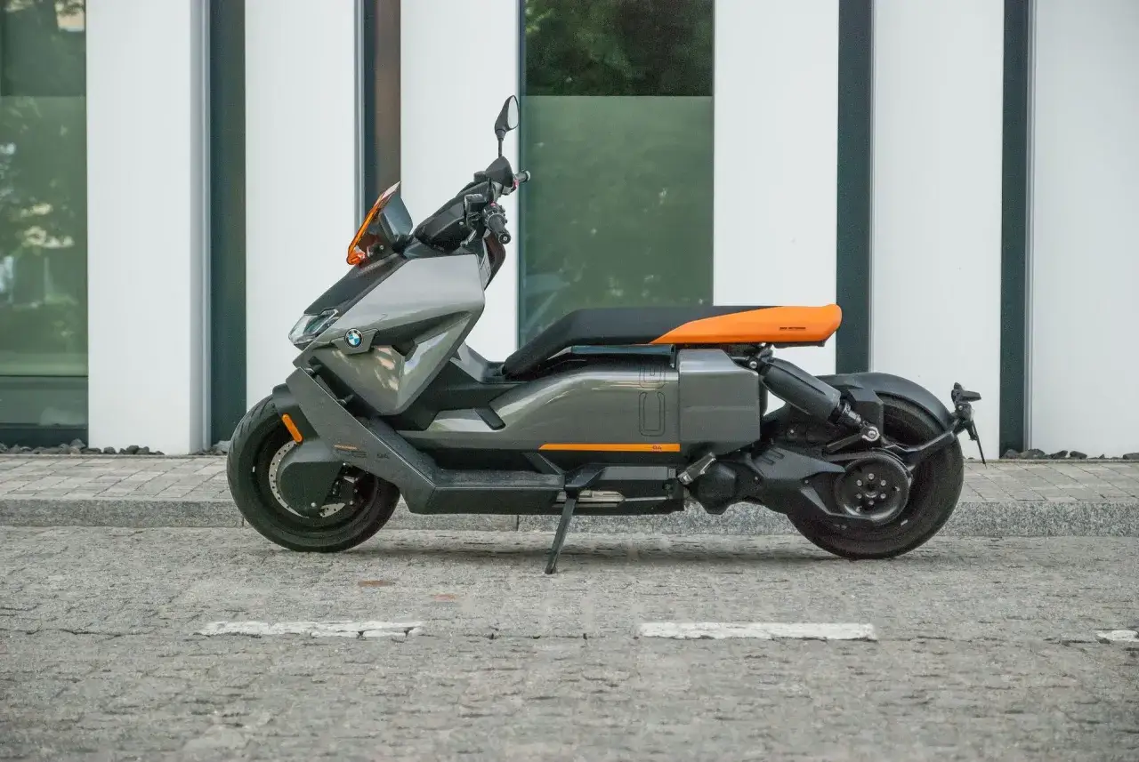 Nowoczesny skuter elektryczny BMW CE 04 w szarym kolorze z pomarańczowymi akcentami stoi na brukowanej ulicy przed minimalistycznym budynkiem.