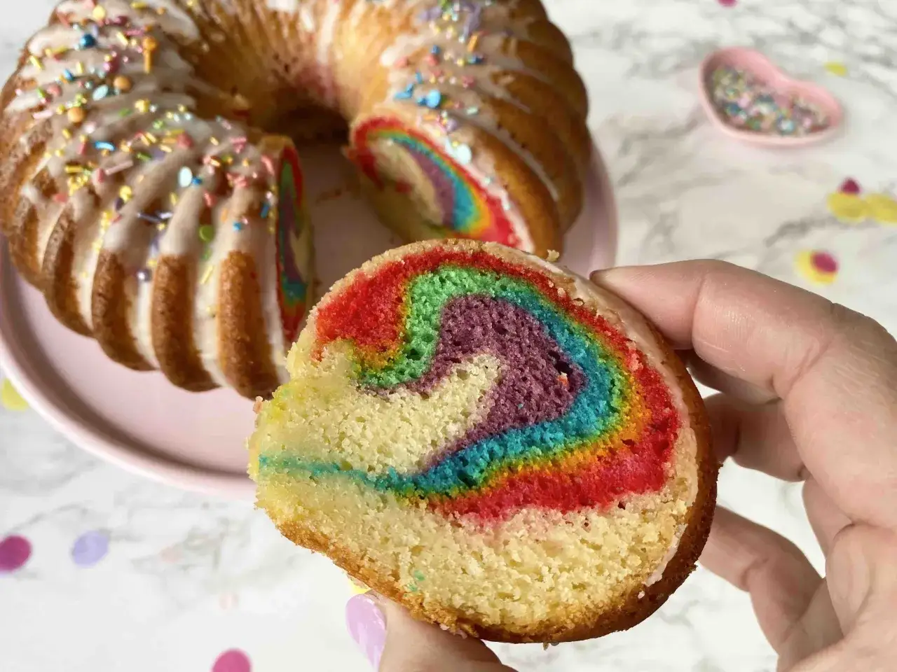 Ein Stück eines bunten **Regenbogenkuchens** mit Zuckerguss und Streuseln wird von einer Hand gehalten. Der Kuchen hat leuchtende Farben im Inneren.