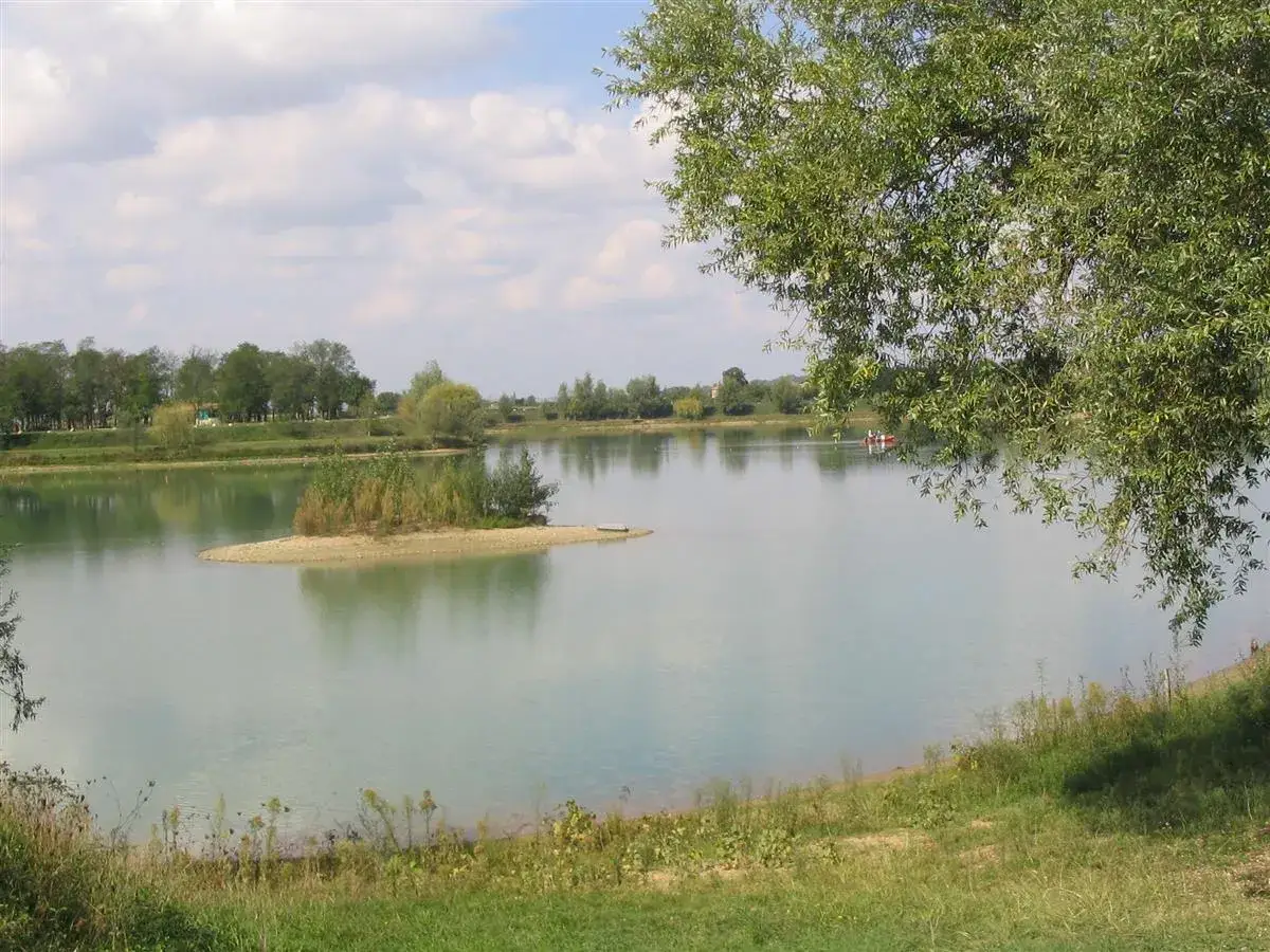 Le lac de Beaupuy, paisible, avec une petite île verdoyante au centre. Un bateau rouge navigue doucement sur l'eau calme.