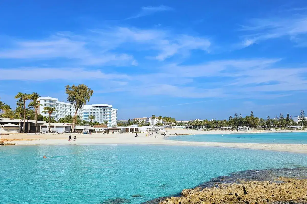 Türkisblaues Wasser und weißer Sand an einem **zypern strand**. Palmen und Hotels säumen die Küste unter blauem Himmel.