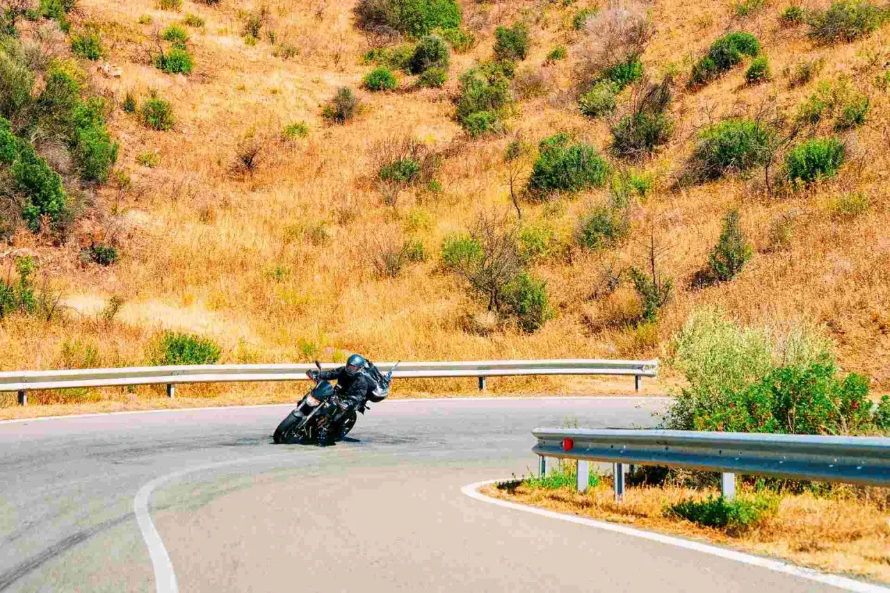 Motociclista in curva su strada panoramica, ideale per itinerari moto Sardegna 7 giorni. Paesaggio arido e vegetazione mediterranea.