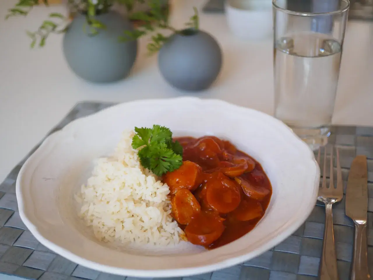 En tallrik med ris och korvgryta, garnerad med persilja. Ett glas vatten och bestick står bredvid. Ett enkelt korvgryta recept.