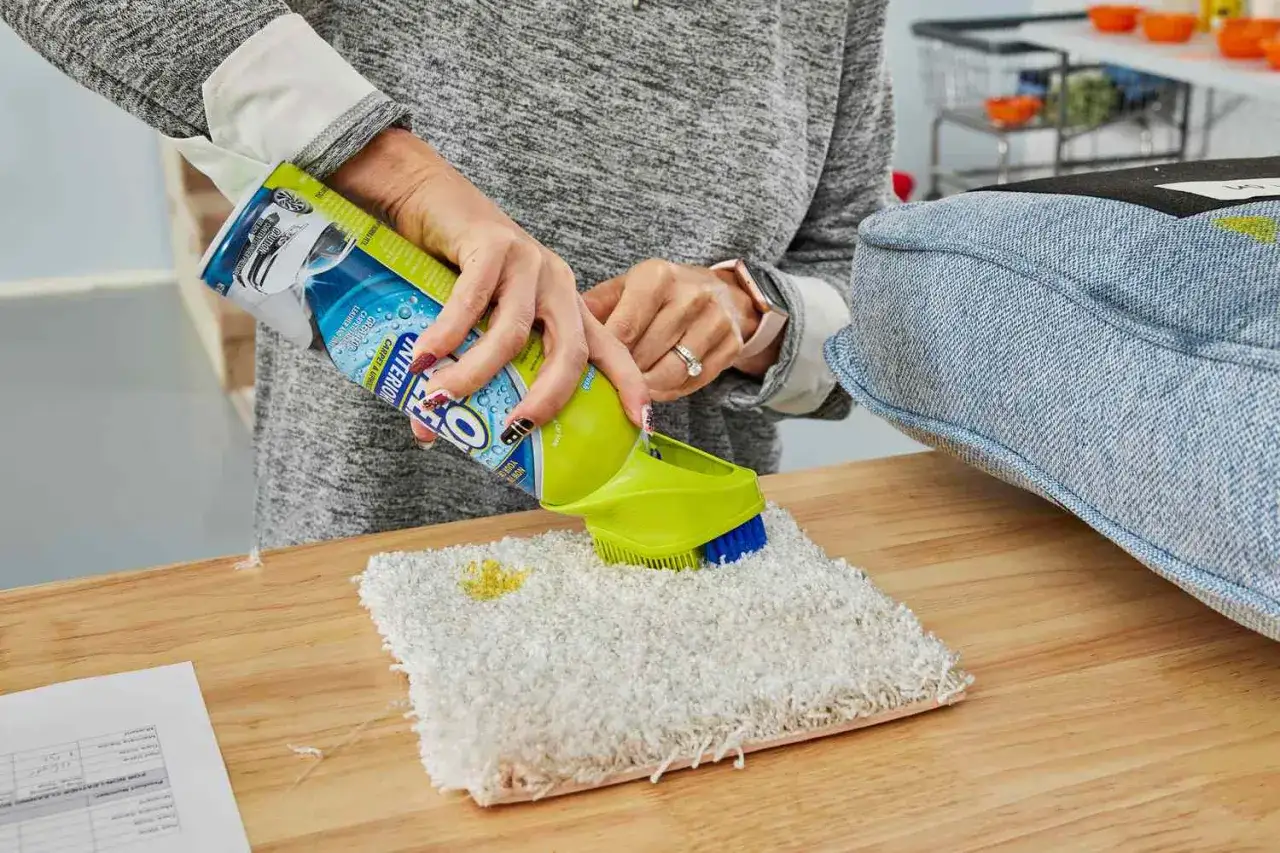 Demonstrating how to clean a couch, a person sprays carpet cleaner onto a swatch and uses a brush.