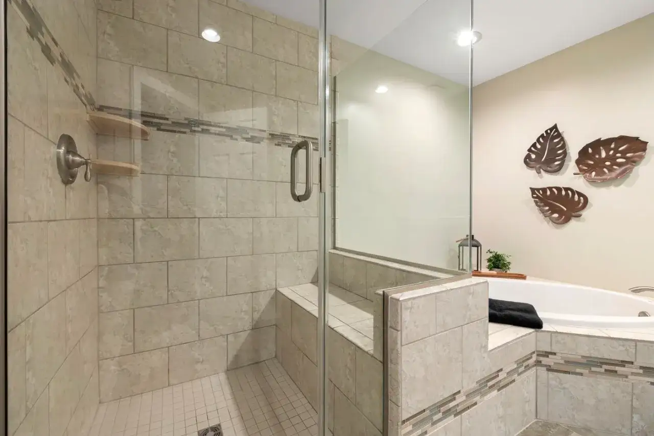 Duchas con murete y cristal, con banco integrado y azulejos de piedra. Ba&ntilde;era adyacente con decoraci&oacute;n de hojas.