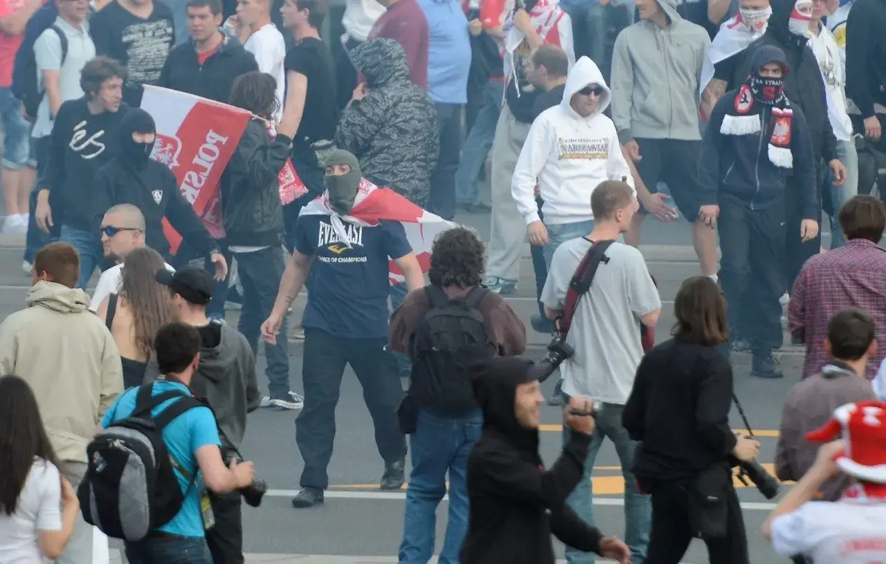 Tłum ludzi, niektórzy w kominiarkach, z polską flagą. Widać napięcie, czy to chuligan czy kibic?