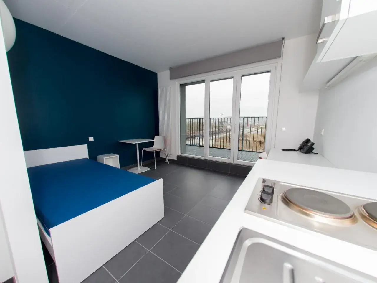 Chambre moderne pour logement étudiant Luxembourg : lit bleu, bureau, kitchenette et balcon avec vue.