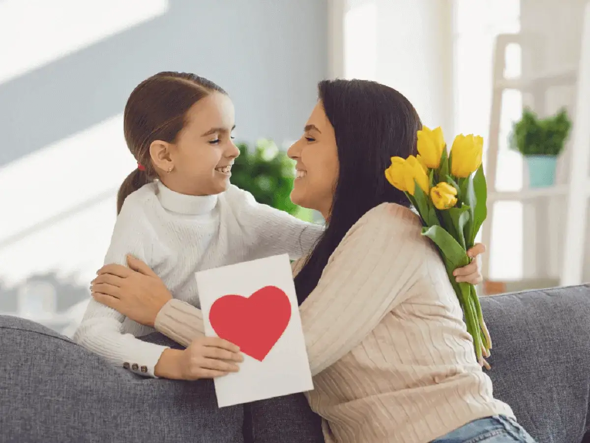Uśmiechnięta mama i c&oacute;rka świętują Dzień Matki. Dziewczynka daje mamie kartkę z sercem i bukiet ż&oacute;łtych tulipan&oacute;w.