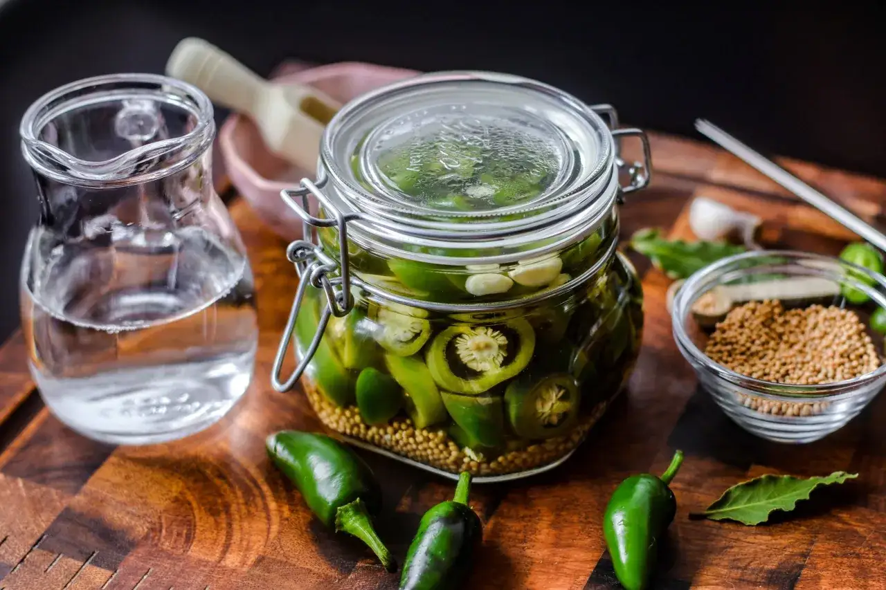 Einmachglas mit geschnittenen Jalapeños, Knoblauch und Senfkörnern, bereit zum Jalapenos einlegen. Daneben ein Krug mit Flüssigkeit.