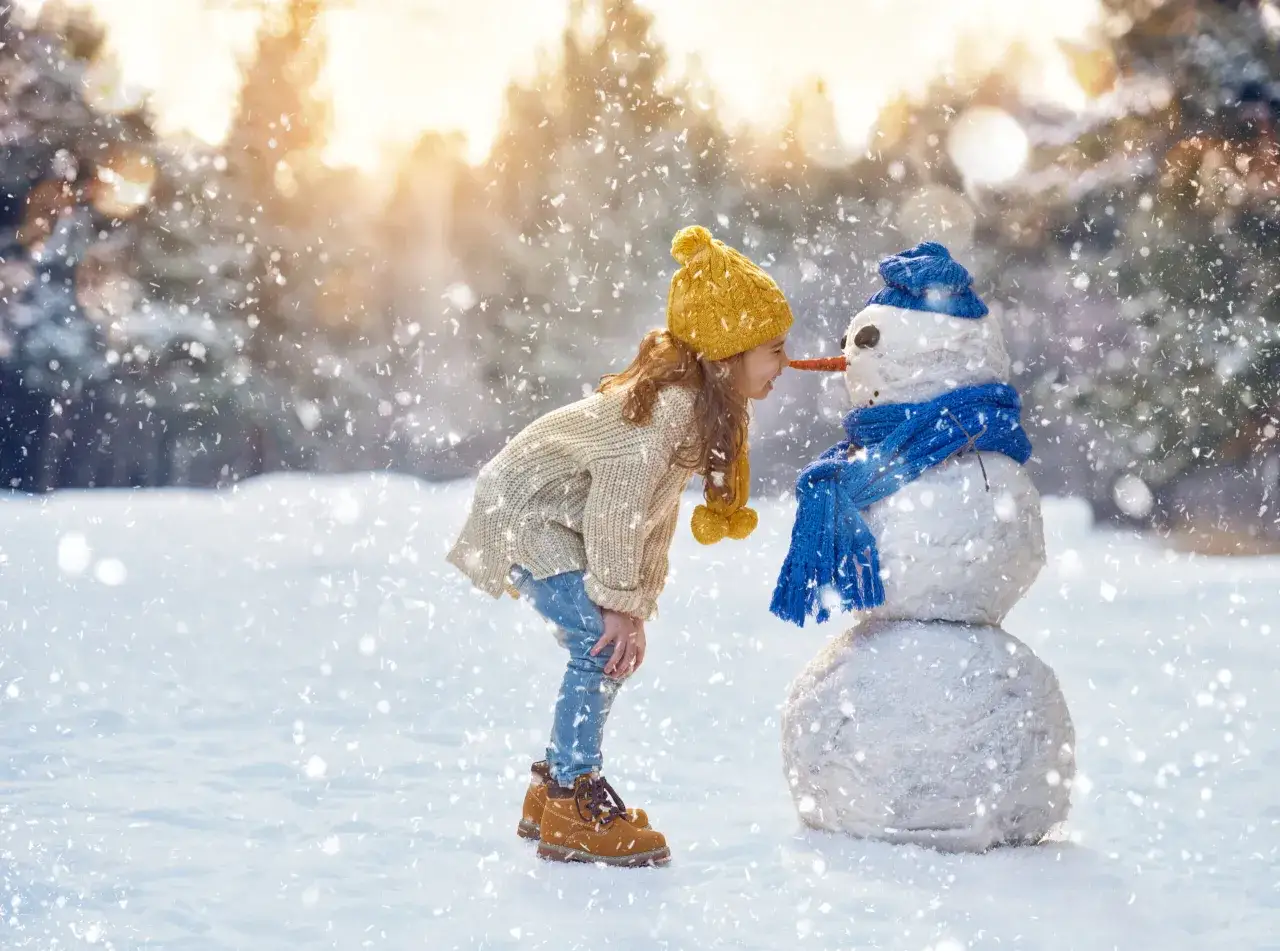 Dziewczynka w żółtej czapce i swetrze pochyla się do bałwanka z marchewkowym nosem. Idealne miejsce, gdzie w góry z dziećmi zimą.