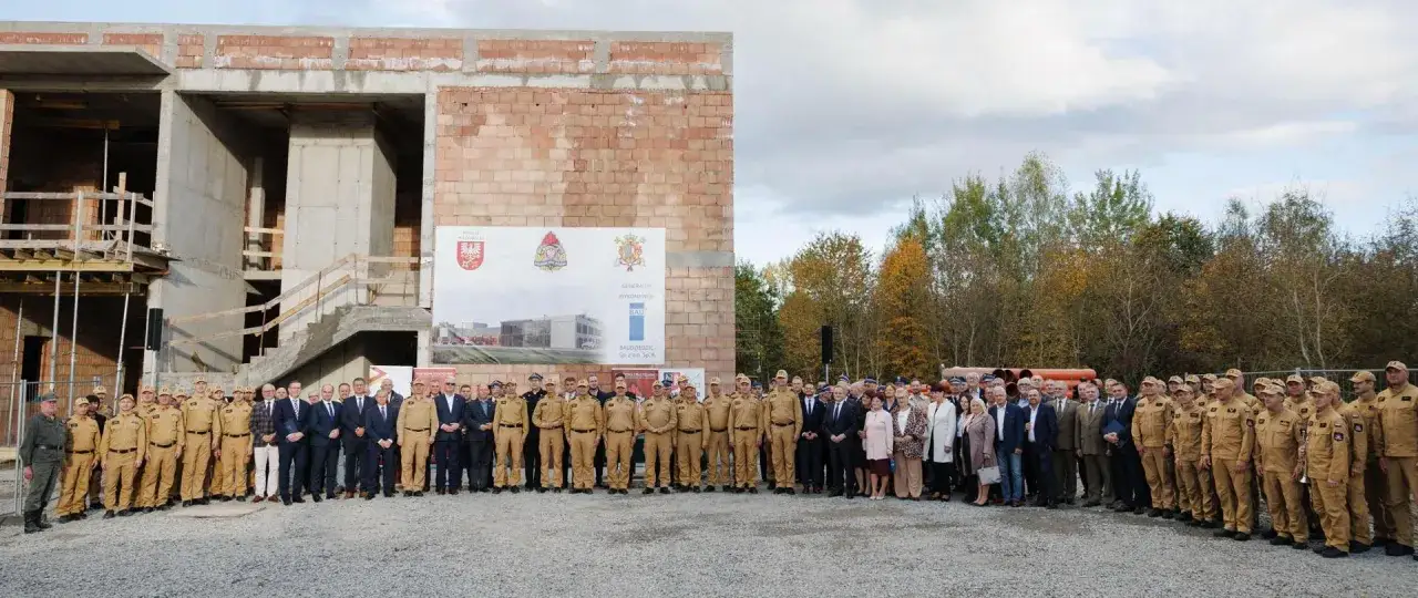 Grupa ludzi pozuje do zdjęcia przed budynkiem w budowie. W tle widoczny jest baner z napisem 