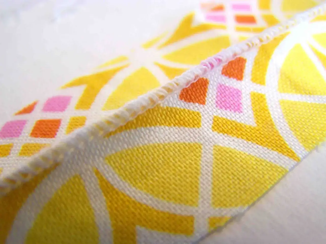 Close-up of a fabric with a yellow, white, pink, and orange geometric pattern. The edge is finished with a decorative stitch, resembling a pintuck.