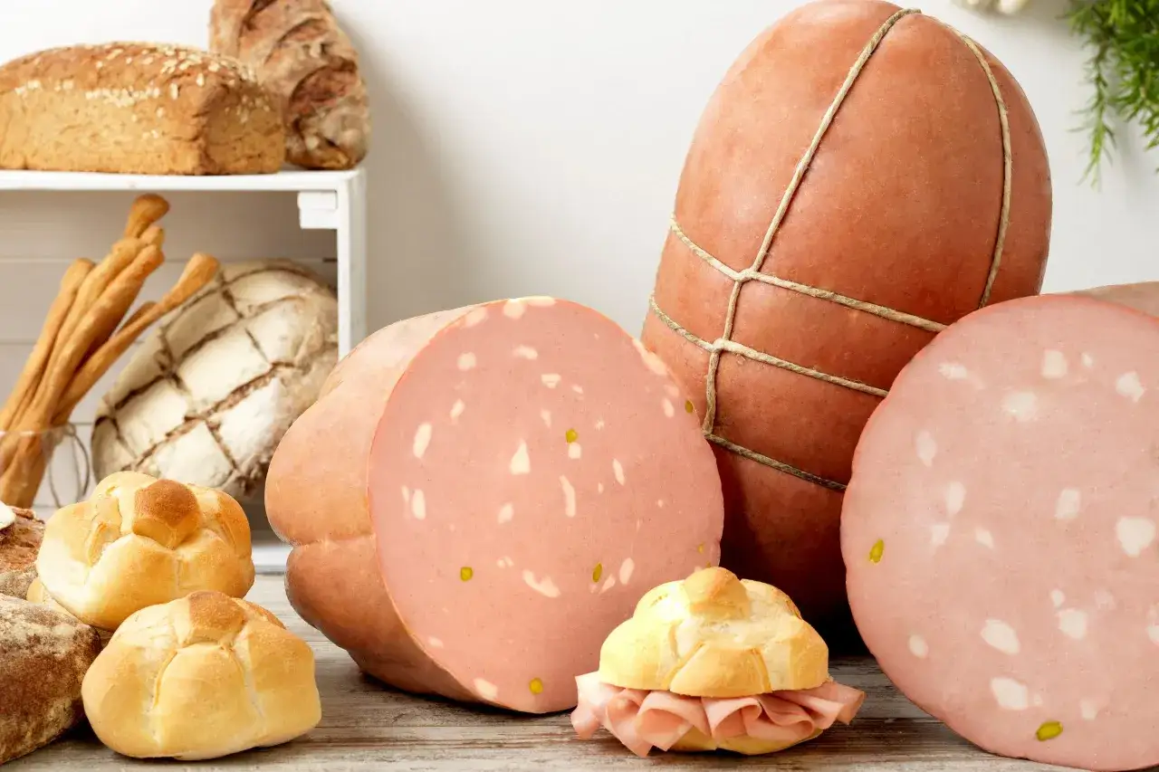 La mortadella è cotta, con fette rosa e bianche, accanto a pane fresco e grissini.