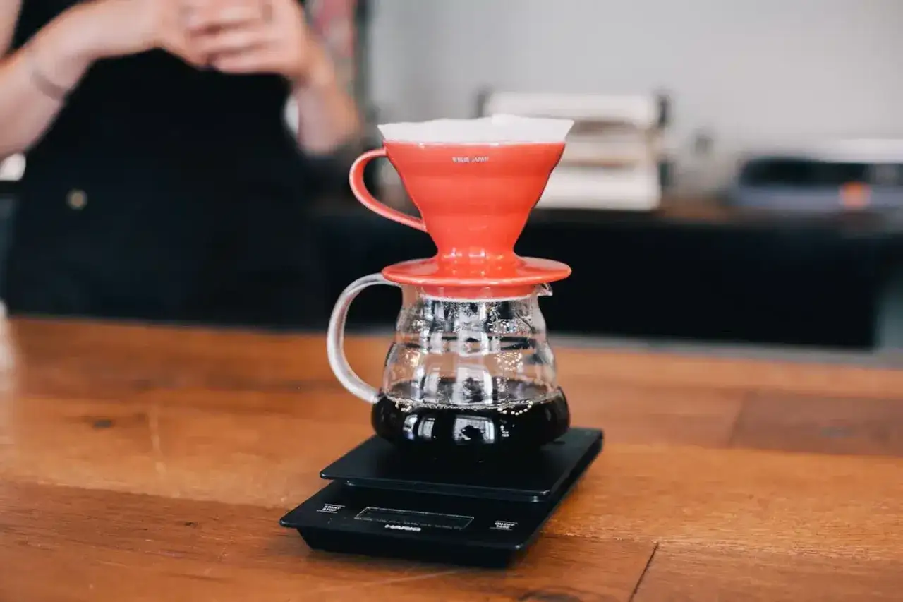 Frisch gebrühter filterkaffee mit milchschaum tropft in eine Glaskaraffe. Eine Person im Hintergrund bereitet den Kaffee zu.