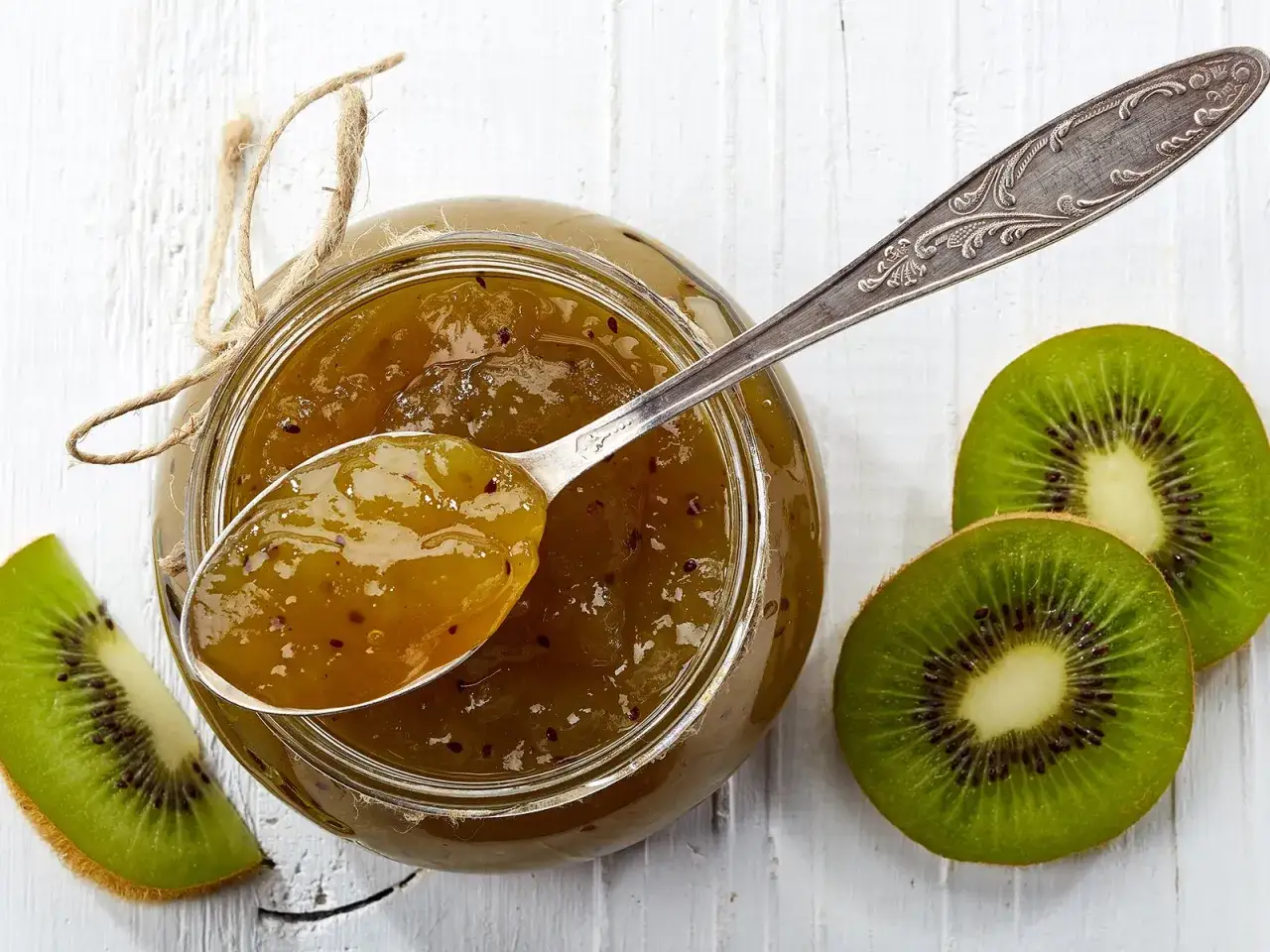 Glas mit leckerer kiwi-marmelade mit schale, daneben frische Kiwischeiben und ein Löffel voll Marmelade.