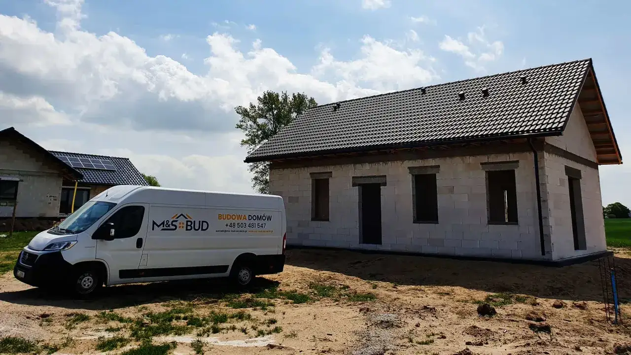 Biały van budowlany M&S BUD stoi obok domu w budowie z szarym dachem i pustymi otworami okiennymi.