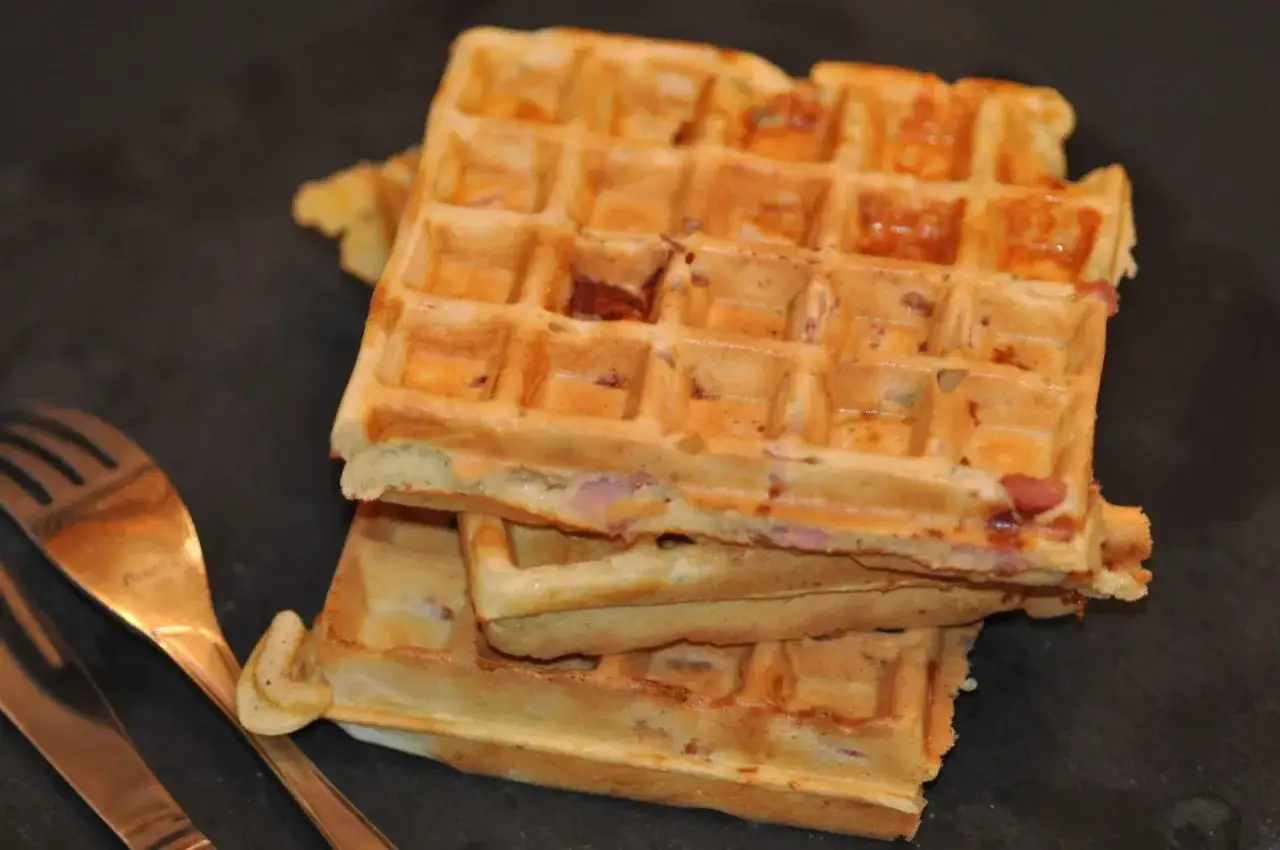 Trois gaufres dor&eacute;es empil&eacute;es, garnies de jambon et d'herbes, pr&ecirc;tes &agrave; &ecirc;tre d&eacute;gust&eacute;es. Une fourchette et un couteau en cuivre sont &agrave; gauche.