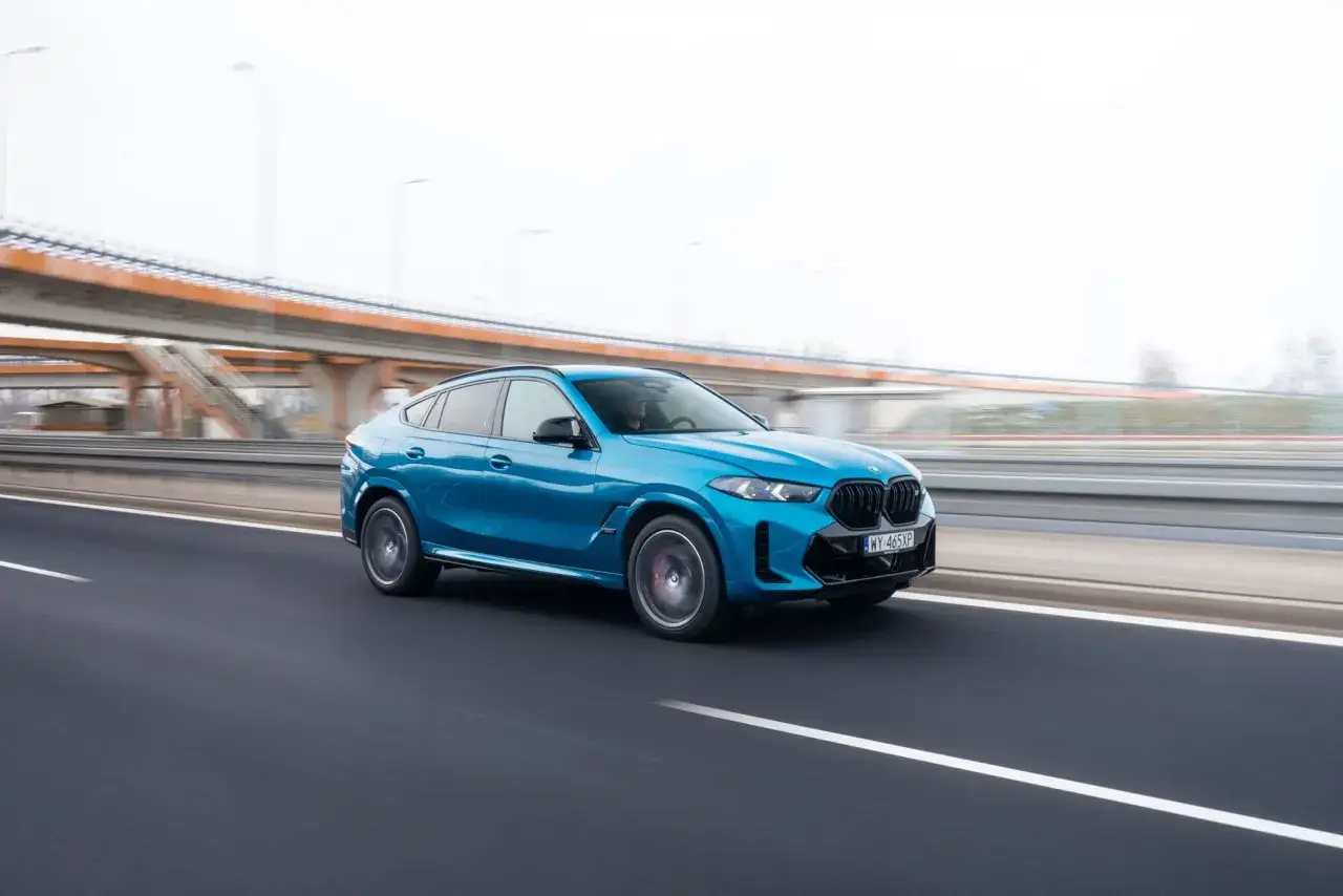 Niebieskie BMW X6M pędzi autostradą, mijając pomarańczową estakadę.