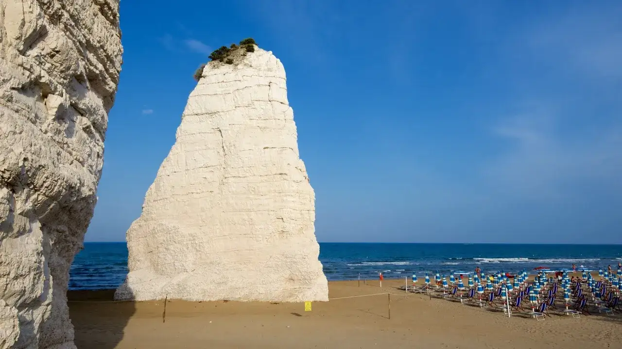 Scogliera bianca a picco sul mare blu, con ombrelloni e lettini sulla spiaggia del comune di Vieste.