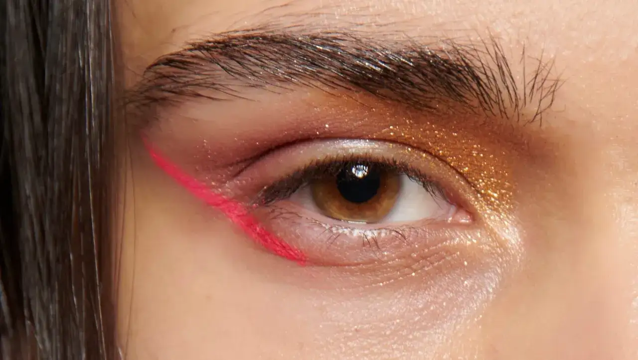 Occhio con trucco audace: glitter dorati sulla palpebra e un audace reverse eyeliner fucsia sotto l'occhio.