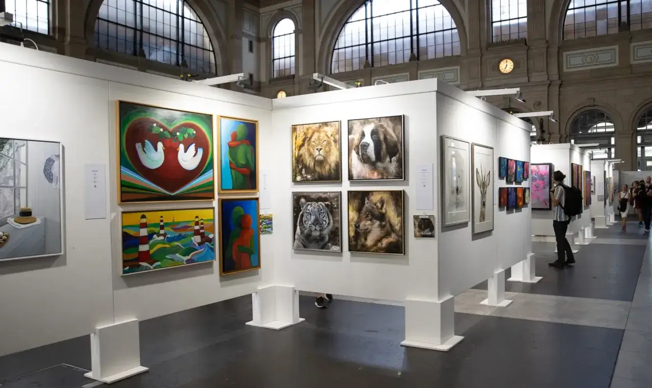 Exhibici&oacute;n de arte con cuadros de animales y paisajes, personas observando las obras en estas ferias de arte.