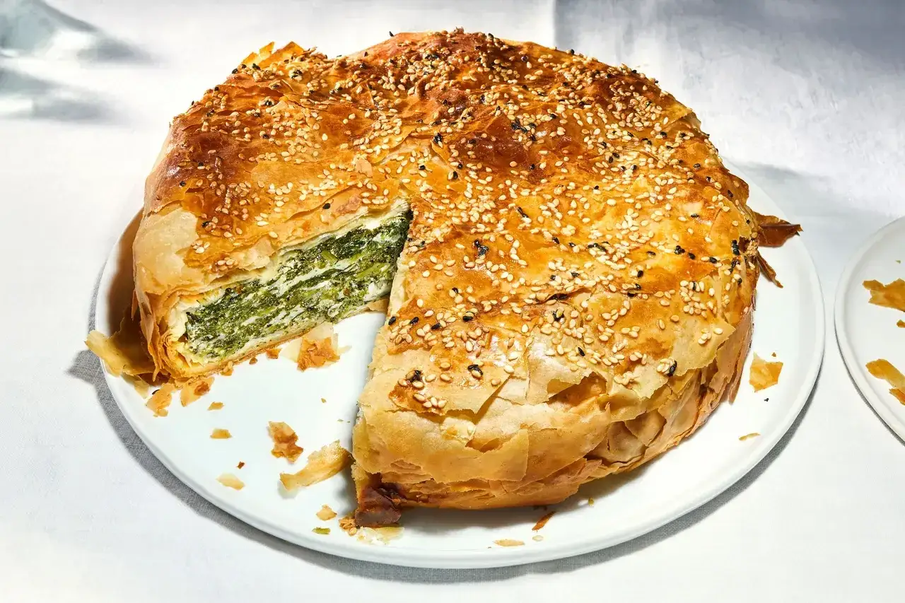 Goldbrauner griechischer Spinatkuchen mit Sesam bestreut, ein St&uuml;ck herausgeschnitten, das die gr&uuml;ne F&uuml;llung zeigt.