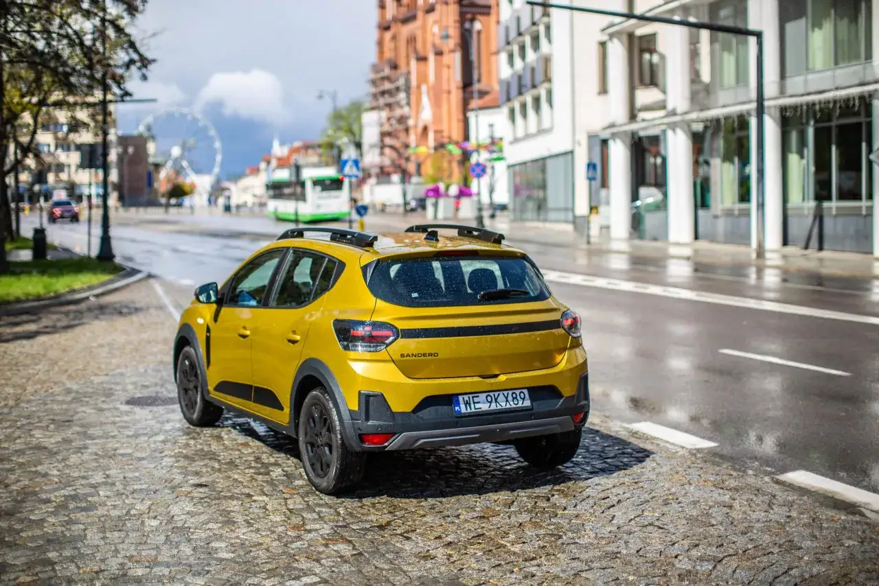 Żółty Dacia Sandero na mokrej, brukowanej ulicy, w tle budynki i diabelski młyn.
