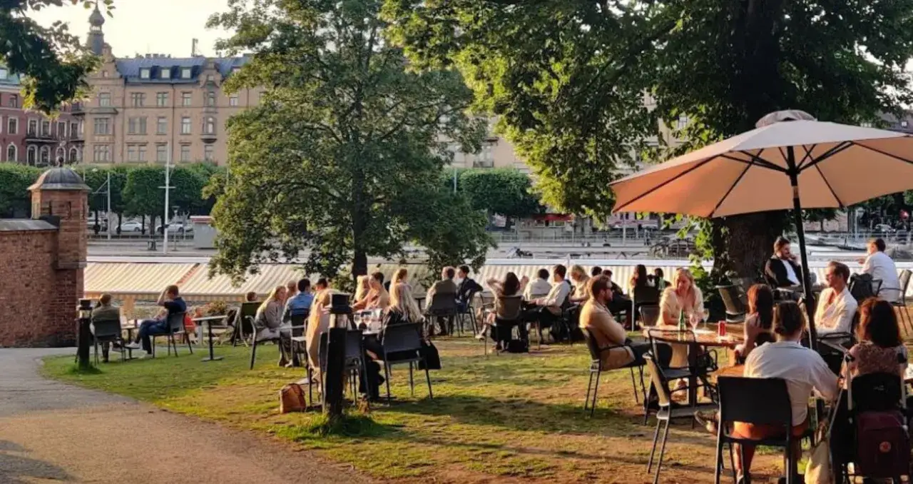 Människor njuter av en solig dag på uteserveringar i Stockholm, med grönska och vackra byggnader i bakgrunden.