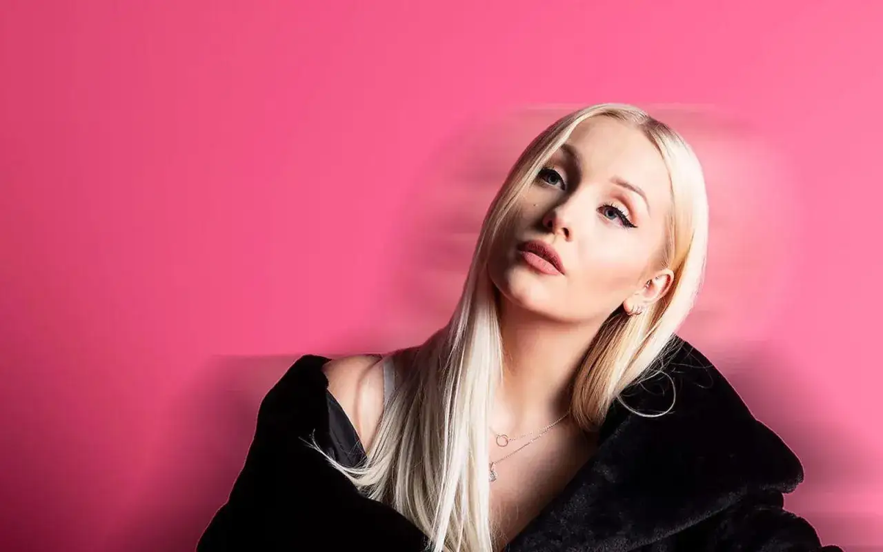 Alexandra Salomonsson med blont h&aring;r och en svart p&auml;lsjacka mot en rosa bakgrund.
