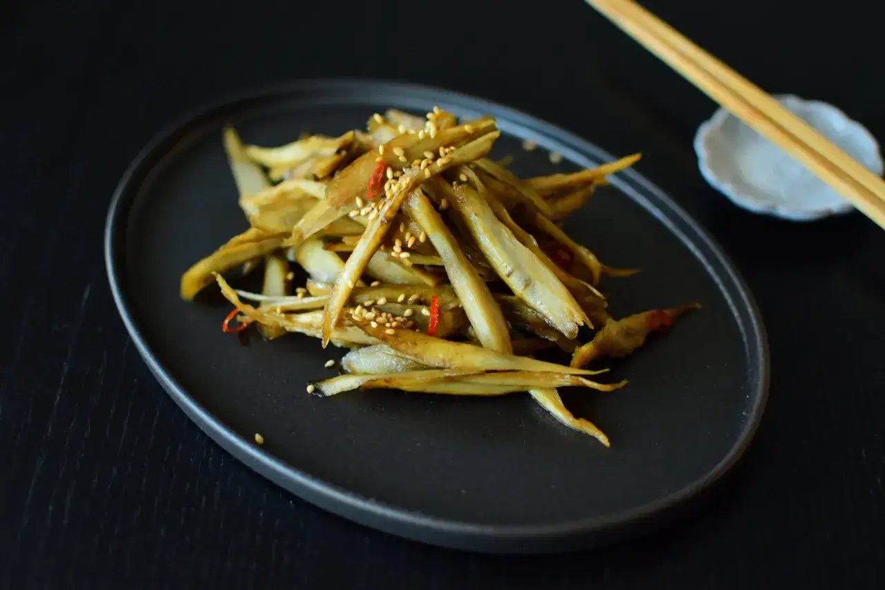 A pile of braised burdock root, seasoned with sesame seeds and chili flakes, sits on a dark plate.