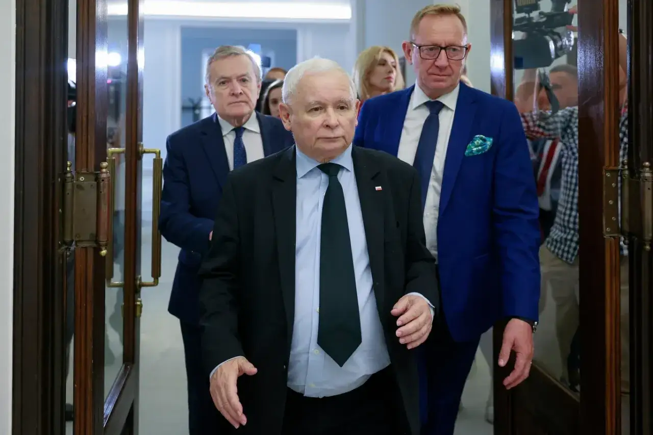 Jarosław Kaczyński, z towarzyszącymi mu mężczyznami, wchodzi przez drzwi. Wzrost i waga polityka są widoczne w jego postawie.