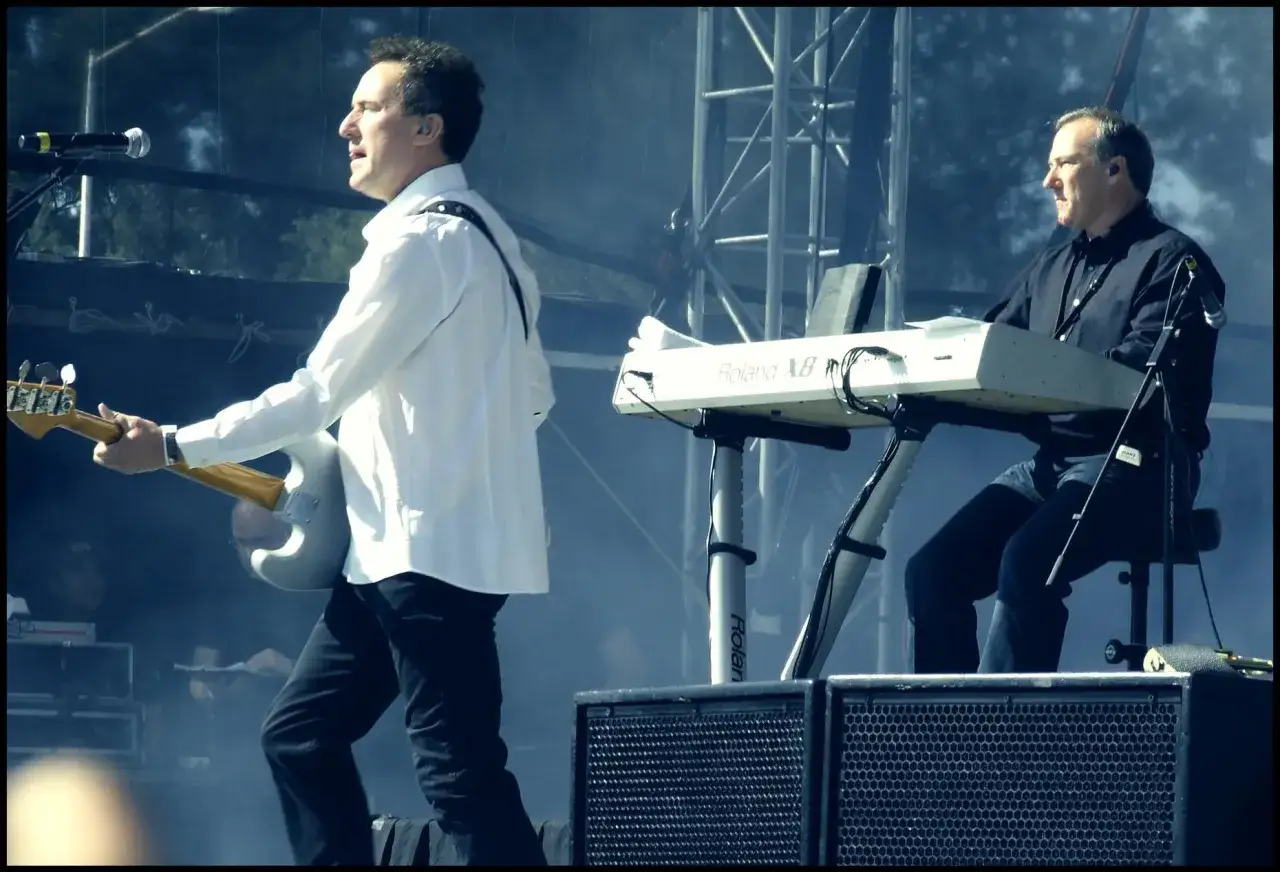 Muzycy OMD grają na żywo. Jeden z nich, w białej koszuli, gra na gitarze basowej, drugi siedzi przy keyboardzie Roland.