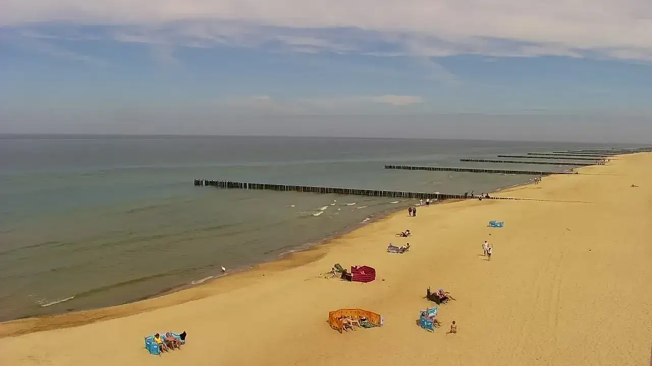 Plaża w Pobierowie z falochronami i spacerującymi ludźmi. Widać parawany plażowe i leżaki, typowe pobierowo atrakcje.