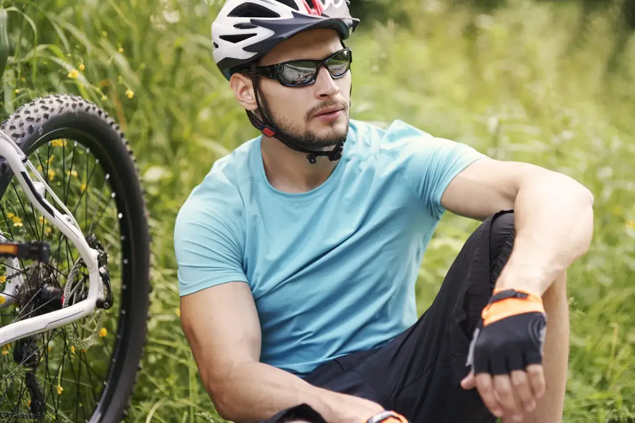 Rowerzysta w kasku i ciemnych okularach, zastanawia się jakie okulary na rower wybrać, siedząc obok swojego roweru w trawie.
