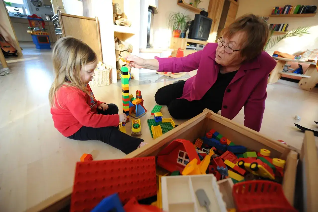 Eine Frau wird zur Leihoma und spielt mit einem Kind im Wohnzimmer mit bunten Bausteinen.