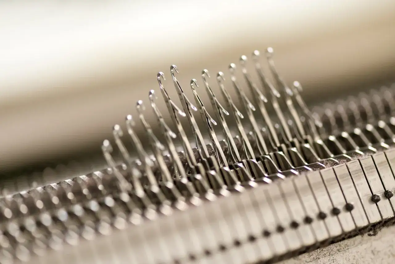 D&eacute;tail d'une machine &agrave; tricoter, montrant les aiguilles m&eacute;talliques align&eacute;es, pr&ecirc;tes &agrave; former des mailles. La jauge maille est pr&eacute;cise.