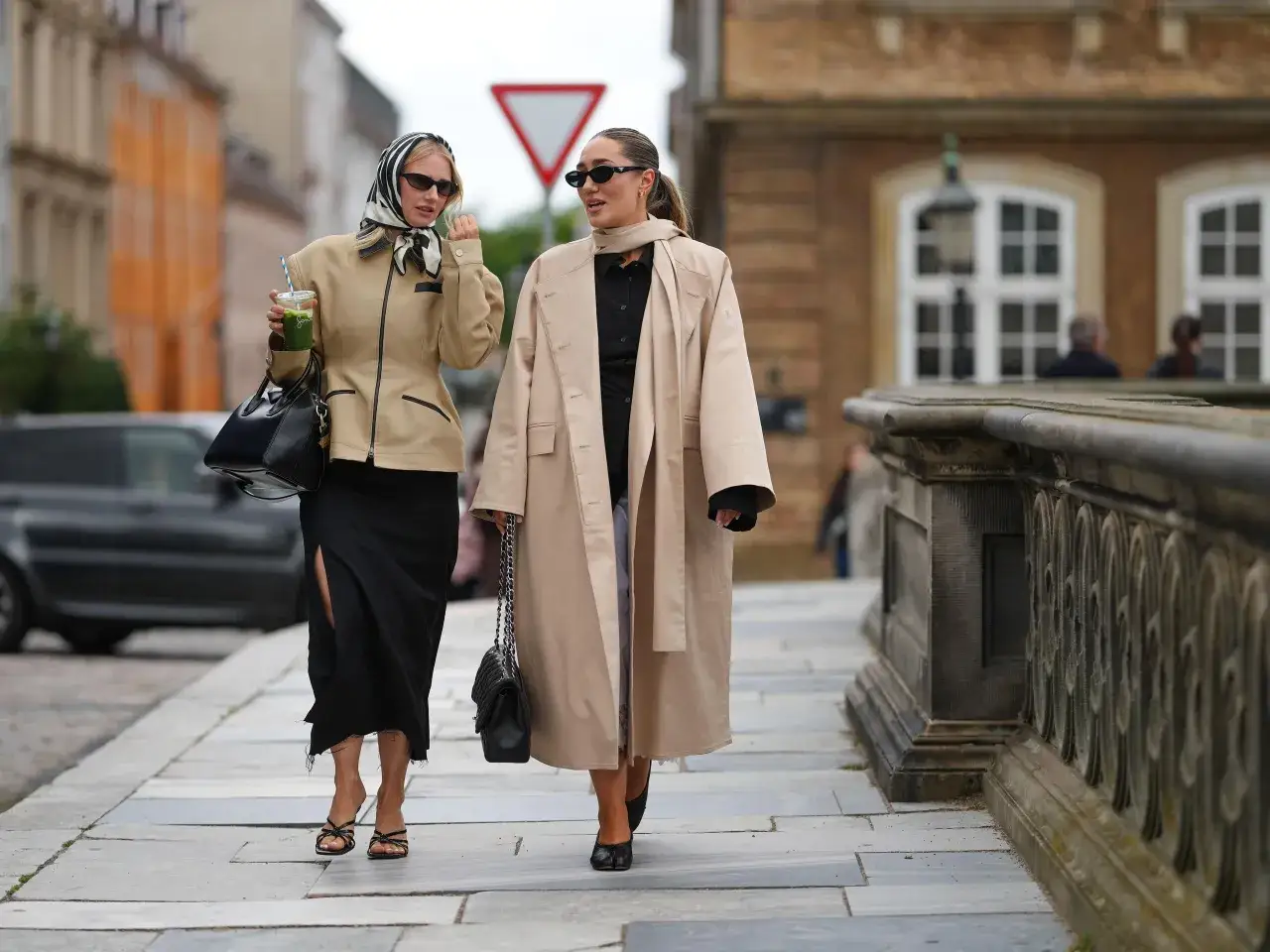 Dos mujeres pasean con estilo. Una lleva una chaqueta corta y falda negra, la otra un gabán largo, como se llaman las chaquetas largas.