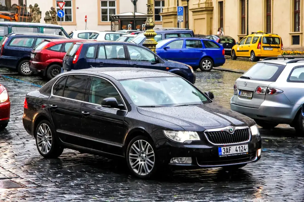 Czarny sedan na mokrej, brukowanej ulicy. Obok inne samochody, w tym żółta taksówka i niebieski hatchback.