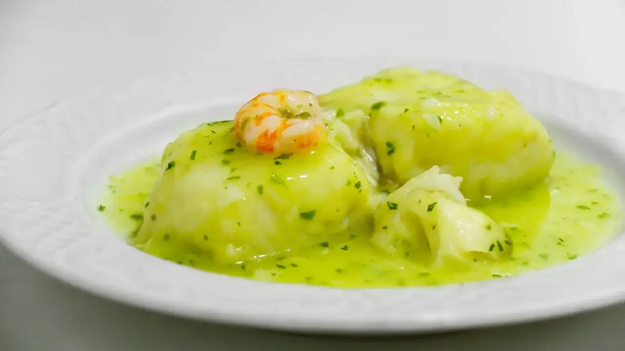 Deliciosa merluza en salsa verde, con un toque de gamba, servida en un plato blanco.
