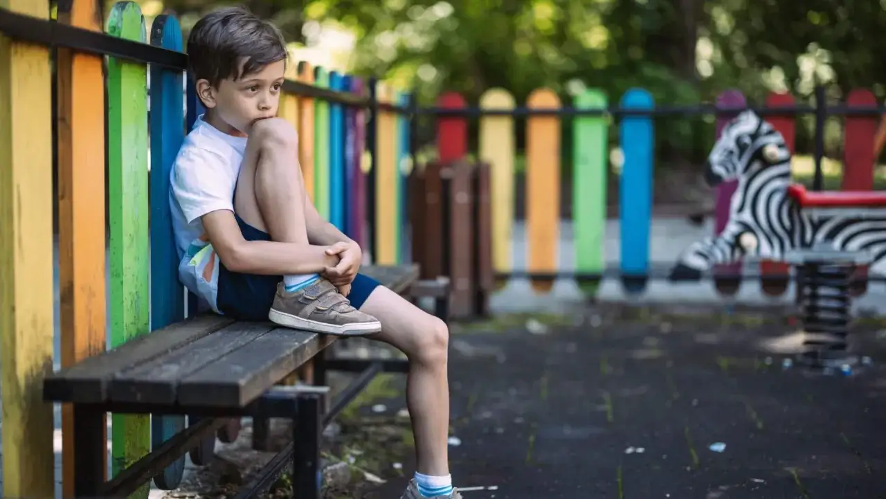Un niño de 4-5 años muestra timidez en el parque, sentado solo en un banco, observando el juego.