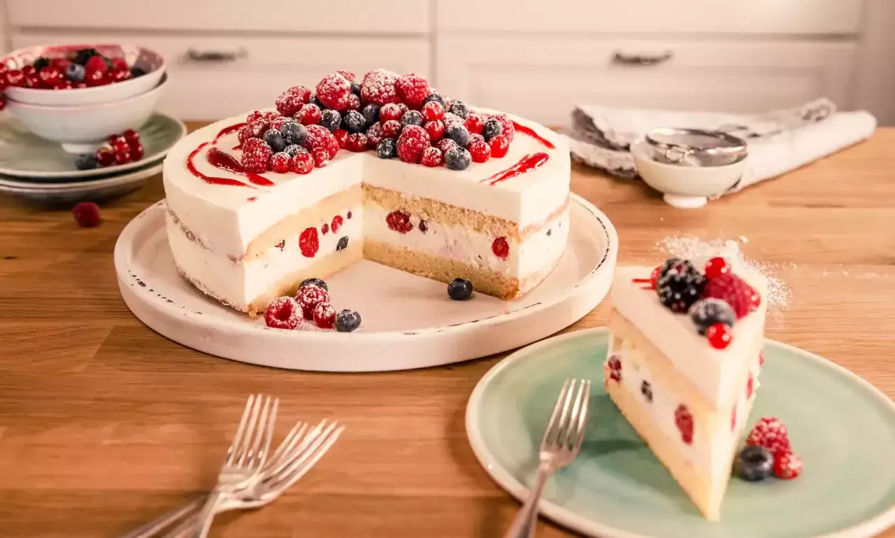 Eine leckere Torte mit Sahne und frischen Beeren, wie Himbeeren und Blaubeeren, verziert. Ein St&uuml;ck ist bereits angeschnitten und auf einem Teller angerichtet.
