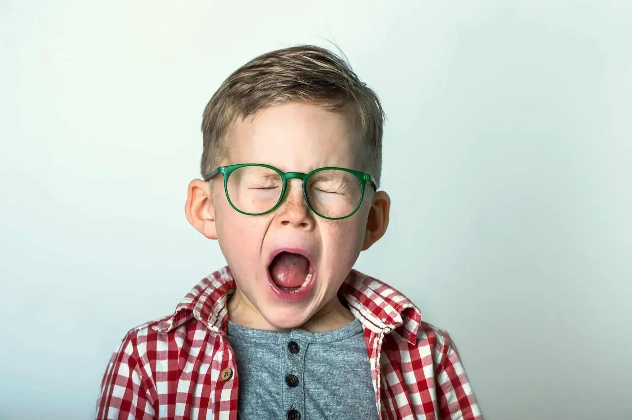 Niño con gafas verdes y camisa a cuadros, con la boca abierta en un bostezo o grito. Podría representar la intensidad de las emociones en el síndrome de Tourette en niños.