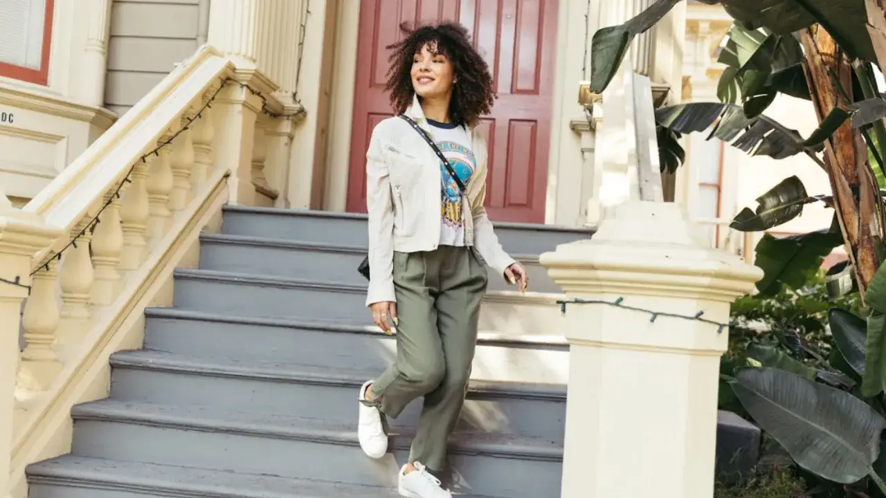 Frau in **smart casual** Kleidung steigt eine Treppe hinunter. Sie trägt eine weiße Jacke, ein bedrucktes T-Shirt und grüne Hosen.