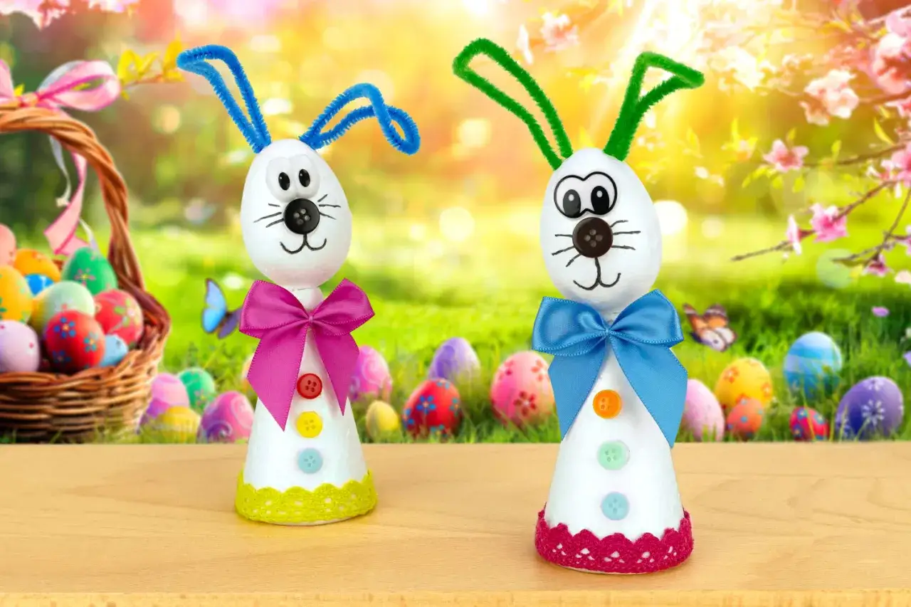 Deux adorables lapins de P&acirc;ques bricolage, faits maison, avec des oreilles en chenille et des n&oelig;uds color&eacute;s, posent devant un panier d'&oelig;ufs de P&acirc;ques.