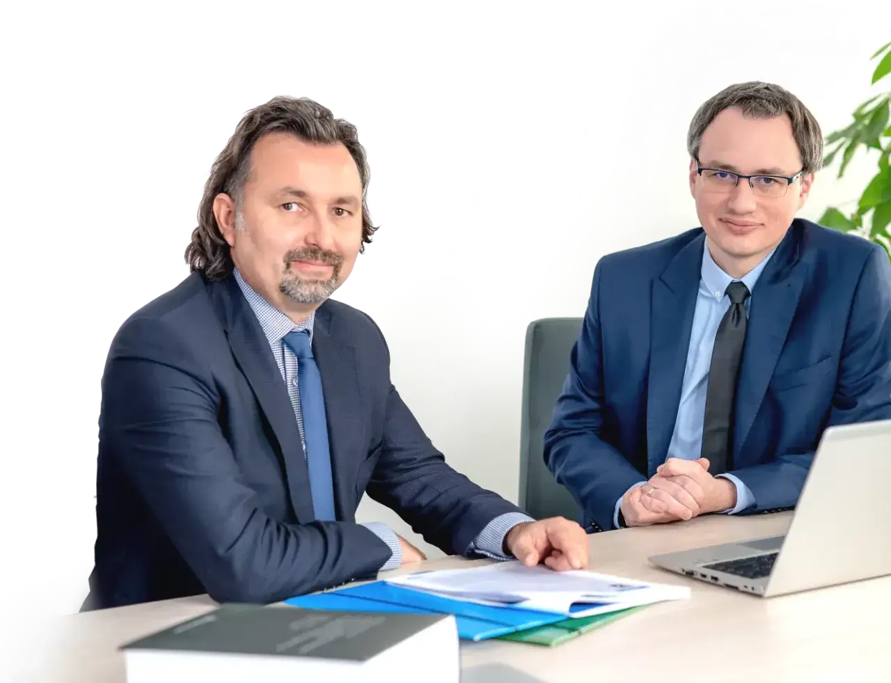Dwóch mężczyzn w garniturach siedzi przy biurku, przed nimi dokumenty i laptop.