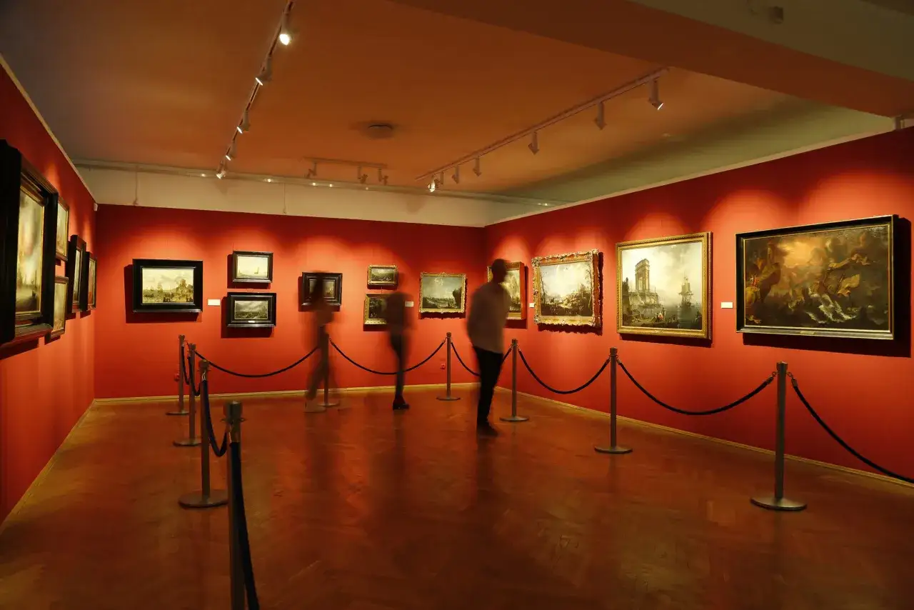 Wnętrze maritime museum gdansk z obrazami marynistycznymi na czerwonych ścianach i rozmytymi postaciami zwiedzających.
