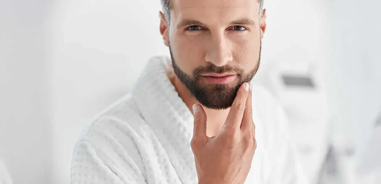 Hombre con barba completa, toc&aacute;ndose la barbilla, con bata blanca.