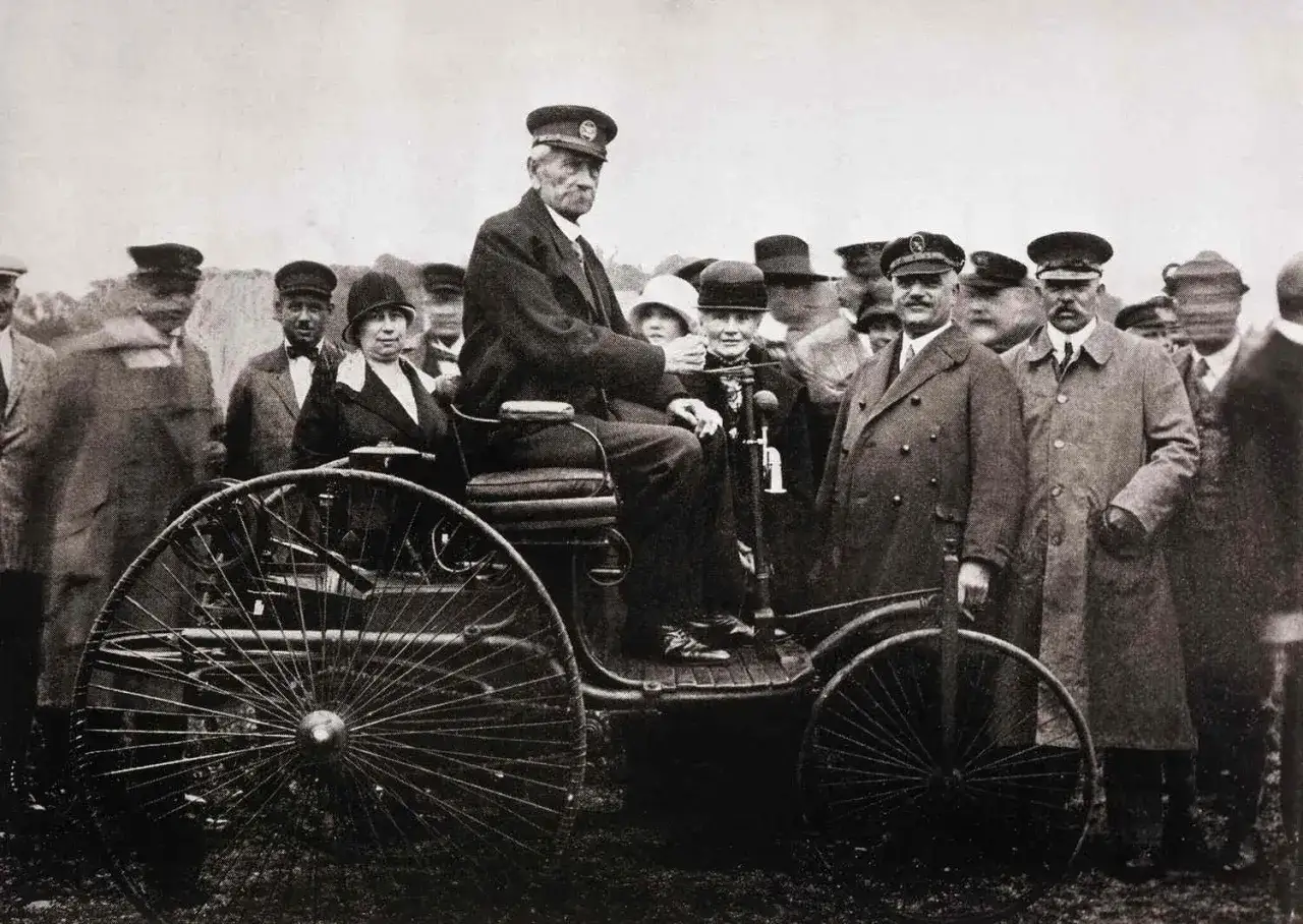 Karl Benz, niemiecki pionier motoryzacji, siedzi za sterami swojego wynalazku, otoczony przez rodzinę i entuzjastów.