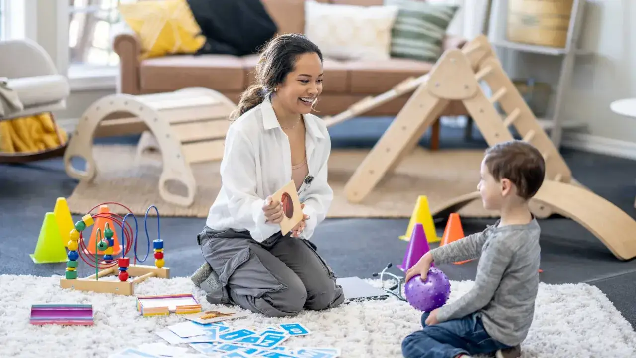 Terapeuta sonriente interactúa con un niño durante una sesión de aba therapy, usando tarjetas y juguetes en un entorno lúdico.